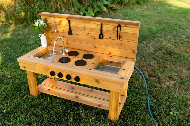 May include: A wooden outdoor play kitchen with a sink, stove, and storage shelf. The kitchen has a water faucet, black burner tops, and hanging utensils. A white vase with flowers sits on the counter. The play kitchen is on a green lawn.