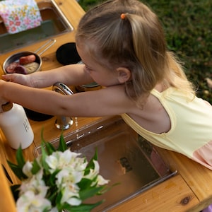 May include: A child plays at a wooden outdoor play kitchen. The kitchen includes a stainless steel sink, a faucet, and various toy cooking accessories. A white bottle with the letters "HI" is on the counter, and a bouquet of white flowers is in the foreground.
