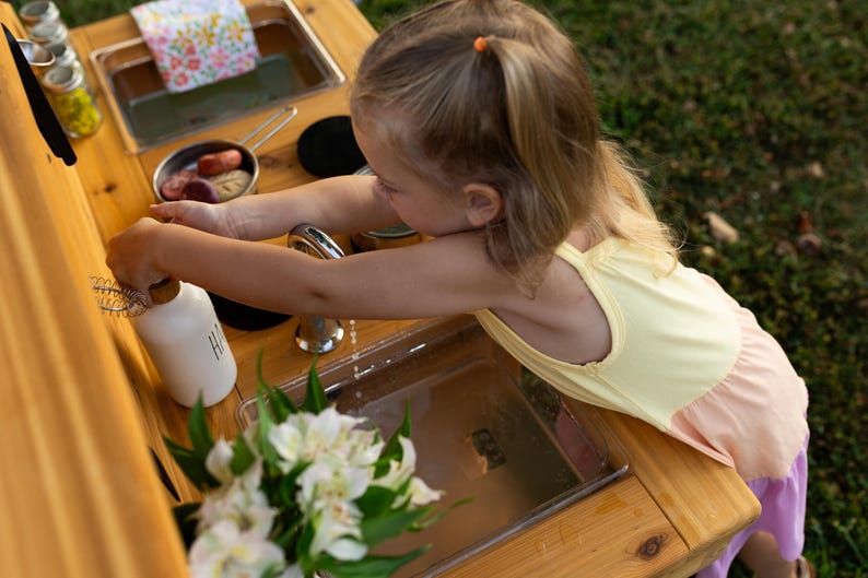 May include: A child plays at a wooden outdoor play kitchen. The kitchen includes a stainless steel sink, a faucet, and various toy cooking accessories. A white bottle with the letters "HI" is on the counter, and a bouquet of white flowers is in the foreground.