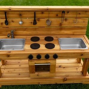 May include: A wooden play kitchen with a natural finish, featuring two stainless steel sinks, four black stovetop burners, and a small oven. It includes hanging utensils and is designed for outdoor play.