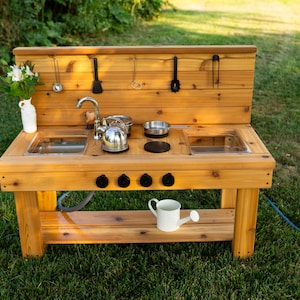 May include: A wooden outdoor play kitchen with a sink, stove, and storage shelf. It includes a metal kettle, bowls, and utensils. The play kitchen is made of light-colored wood and sits on a grassy lawn.