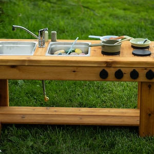 Peut inclure: Une cuisine extérieure en bois avec un évier en acier inoxydable, un robinet et une plaque de cuisson. La cuisine a une étagère inférieure et quatre boutons noirs. Les accessoires ne sont pas inclus. La cuisine est posée sur une pelouse verte.