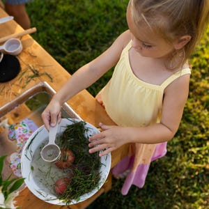 May include: A young child in a yellow top and pink skirt plays with water and natural elements in a white bowl. The scene includes a wooden table with play kitchen accessories, a silver faucet, and green grass.
