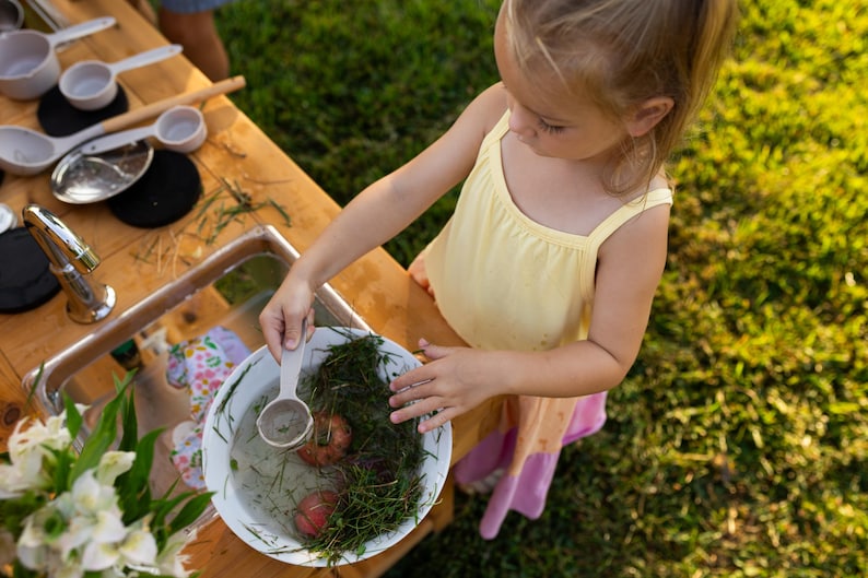 May include: A young child in a yellow top and pink skirt plays with water and natural elements in a white bowl. The scene includes a wooden table with play kitchen accessories, a silver faucet, and green grass.