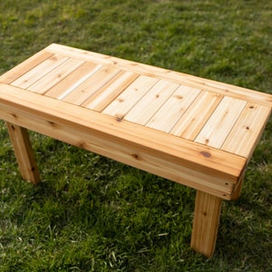 May include: A rectangular wooden bench with a natural finish. The bench has a slatted top and four sturdy legs. The wood grain is visible, and the bench is set on a grassy surface. A simple, functional piece of outdoor furniture.