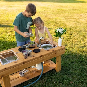 May include: A wooden outdoor mud kitchen featuring a rectangular frame, two stainless steel basins, a metal tap, and a lower storage shelf. The kitchen includes various small metal pots, glass jars, a white pitcher with flowers, and a patterned towel.