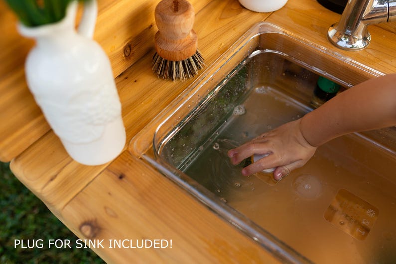 May include: A wooden play sink with a stainless steel basin. A white vase with greenery and a wooden brush are on the wooden surface. The text "PLUG FOR SINK INCLUDED!" is at the bottom of the image.