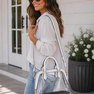 May include: A white and clear handbag with a top handle and a shoulder strap. The bag has gold-colored hardware and a black interior. The bag is being carried by a person wearing a white shirt and blue jeans.