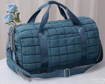 Könnte beinhalten: Türkis gesteppte Reisetasche mit einem quadratischen Muster. Die Tasche hat zwei obere Griffe und einen abnehmbaren Schultergurt. Zu den Merkmalen gehören eine Vordertasche und silberfarbene Reißverschlüsse. Hergestellt aus weichem, gepolstertem Material.