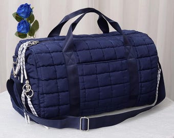 Könnte beinhalten: Eine marineblaue, gesteppte Reisetasche mit einem rechteckigen Muster. Die Tasche hat einen oberen Griff, einen abnehmbaren Schultergurt und eine Vordertasche. Weiße und silberne Akzente sind an der Seite sichtbar. Die Tasche steht auf einer weißen Oberfläche.