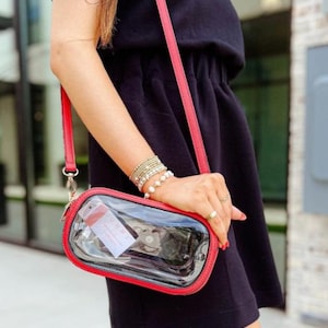May include: A clear crossbody bag with a red trim and strap. The bag is rectangular and has a zipper closure. The strap is thin and adjustable. The bag is being held by a person wearing a black dress.