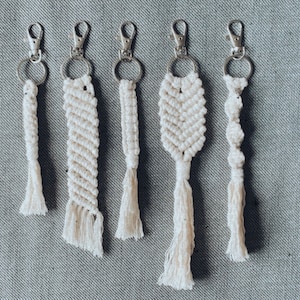 Puede incluir: Seis llaveros de macramé blanco con anillas de plata. Los llaveros están hechos con una variedad de nudos de macramé y tienen borlas en la parte inferior.
