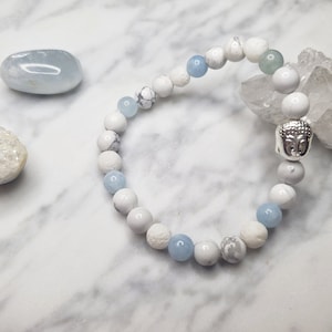 May include: A beaded bracelet featuring white and light blue beads, interspersed with a silver Buddha head charm. The bracelet is displayed with other crystals and stones on a marble surface.