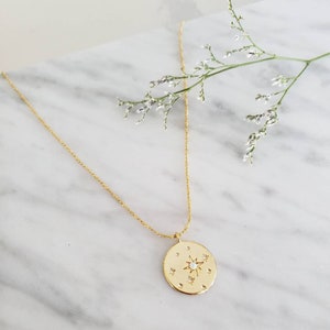 Puede incluir: Un collar de oro con un colgante redondo que presenta un diseño de estrella con un ópalo blanco en el centro y pequeñas piedras transparentes alrededor.