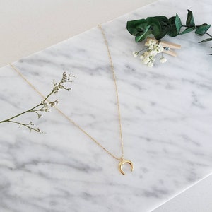 May include: A gold crescent moon pendant necklace on a delicate chain. The necklace is displayed on a white marble surface.
