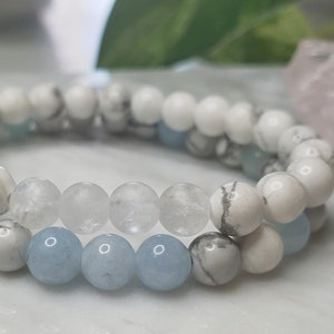 May include: A set of two stretch bracelets featuring round beads. One bracelet consists of white and grey marbled beads with light blue accents. The second bracelet features a combination of white marbled beads, clear translucent beads, and light blue beads.