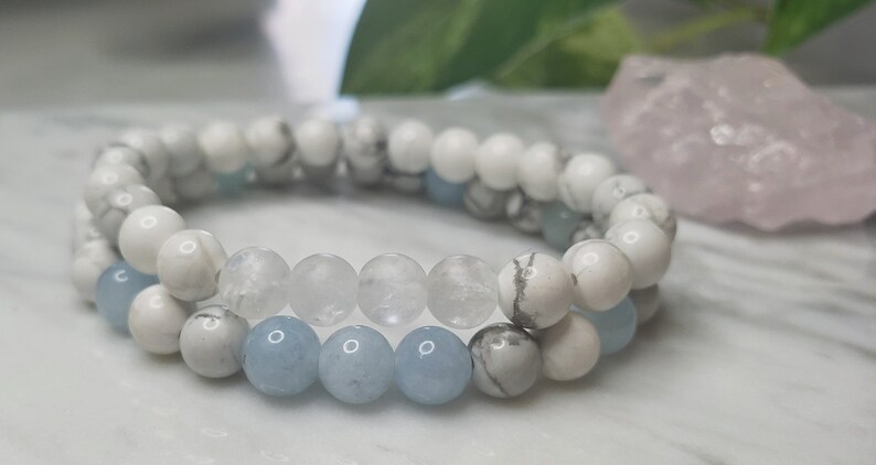 May include: A set of two stretch bracelets featuring round beads. One bracelet consists of white and grey marbled beads with light blue accents. The second bracelet features a combination of white marbled beads, clear translucent beads, and light blue beads.