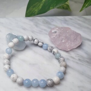 May include: A beaded bracelet featuring alternating round white marbled beads and smooth light blue beads. The bracelet is arranged in a circular shape, showcasing a repeating pattern of stone-like textures and solid translucent blue spheres.