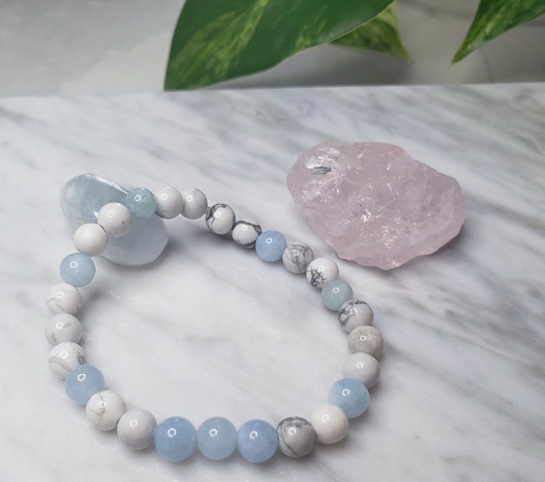May include: A beaded bracelet featuring alternating round white marbled beads and smooth light blue beads. The bracelet is arranged in a circular shape, showcasing a repeating pattern of stone-like textures and solid translucent blue spheres.