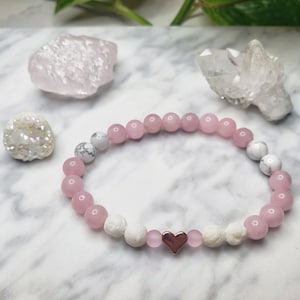 May include: A pink and white beaded bracelet with a rose gold heart charm. The bracelet is made with rose quartz and white howlite beads.