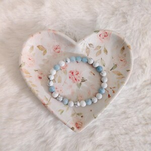 May include: A beaded bracelet featuring alternating round white marble-patterned beads and smooth light blue beads, resting inside a heart-shaped ceramic dish decorated with a delicate pink rose and green leaf floral pattern.