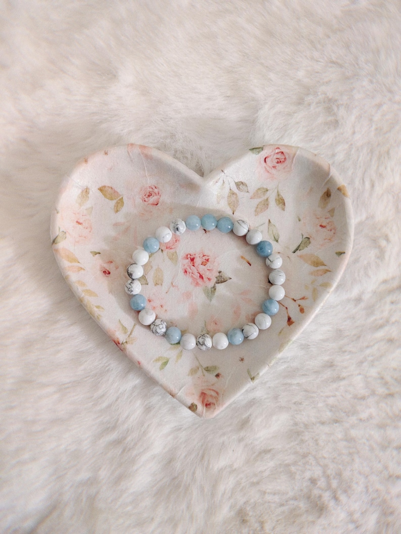 May include: A beaded bracelet featuring alternating round white marble-patterned beads and smooth light blue beads, resting inside a heart-shaped ceramic dish decorated with a delicate pink rose and green leaf floral pattern.