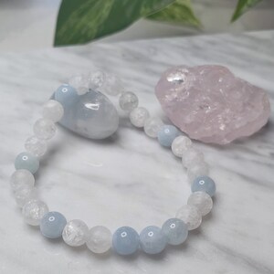 May include: A light blue and white beaded bracelet with clear quartz and aquamarine stones. The bracelet is on a white marble surface with a pink crystal next to it.
