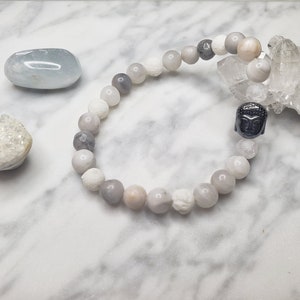 May include: A grey and white beaded bracelet with a black Buddha charm. The bracelet is on a white marble surface with other crystals.