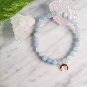 May include: A light blue aquamarine gemstone bracelet with a gold crescent moon charm. The bracelet is on a white marble surface with a pink and white crystal next to it.