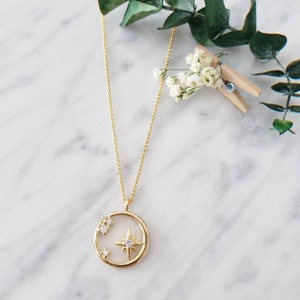 May include: A gold necklace with a circular pendant featuring a starburst design with small clear stones. The pendant is on a delicate chain.