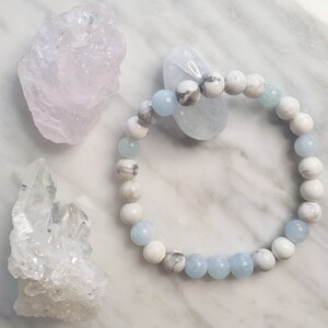 May include: A beaded bracelet featuring alternating round white and grey marbled stone beads and translucent light blue beads. The bracelet is arranged in a circular shape, showcasing a smooth, polished finish on each individual spherical bead.