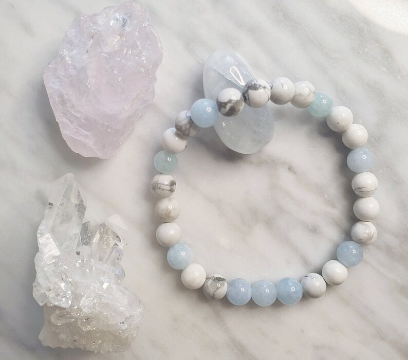 May include: A beaded bracelet featuring alternating round white and grey marbled stone beads and translucent light blue beads. The bracelet is arranged in a circular shape, showcasing a smooth, polished finish on each individual spherical bead.