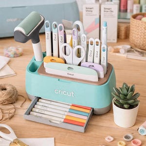 May include: A teal Cricut craft organiser filled with various tools, including pens, scissors, and a roller. The organiser sits on a wooden surface, alongside a small succulent in a white pot and colourful buttons.