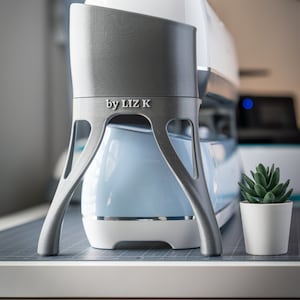 May include: A grey 3D-printed stand with the text "by LIZ K" in front of a white and light blue crafting machine. A small potted succulent is to the right. The stand has a unique, open-legged design.