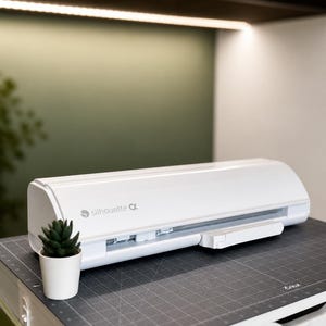 Puede incluir: Una máquina de corte Silhouette Alpha blanca con el nombre de la marca en el lateral. La máquina está sobre una esterilla de corte gris con una pequeña planta suculenta en maceta. El fondo es una pared verde claro y blanca.