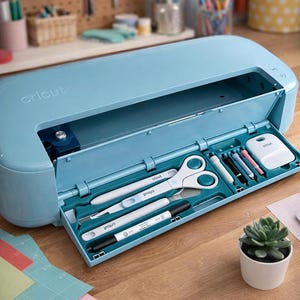 Può includere: Macchina da taglio Cricut azzurra con un vano aperto che rivela vari strumenti per il crafting, tra cui forbici, penne e un raschietto. La macchina è su una superficie di legno, con carta colorata e una piccola pianta grassa in vaso nelle vicinanze.