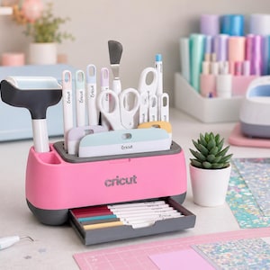 May include: A bright pink Cricut desktop organizer featuring a pull-out drawer containing several pens. The top section holds various crafting tools including a brayer, scissors, and scrapers. The organizer is branded with the Cricut logo on the front.