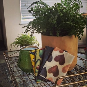 Puede incluir: Una colección de envoltorios de alimentos reutilizables de cera de abeja con varios diseños, incluyendo aguacates y corazones, exhibidos en una rejilla dorada. Un portavelas verde con diseño de hojas y plantas en macetas en el fondo.