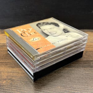 May include: A collection of CDs featuring Patsy Cline. The top CD case is labelled "Patsy Cline Live Volume Two" in orange and white text. The CDs are stacked on a black base. The image shows a portrait of Patsy Cline.