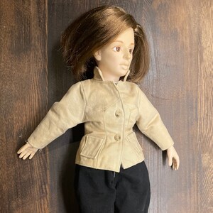 May include: A vintage doll dressed in a tan jacket with three buttons, black pants, and black boots. The doll has brown hair and is posed on a wooden surface. The doll's outfit resembles a riding ensemble.