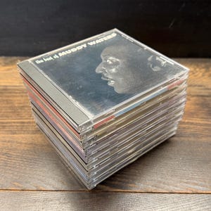 May include: A pile of CDs in clear plastic cases. The top CD case displays a black and white image of a man's profile and the text "the best of Muddy Waters". The CDs are stacked on a wooden surface.