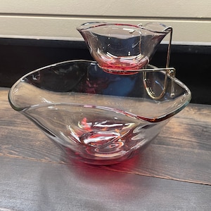 May include: A two-tiered serving set with a clear glass bowl and a smaller bowl suspended above it by a gold-coloured metal frame. The rims of the bowls have a red tint. The set is designed for serving snacks or dips.