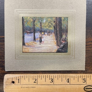 May include: A framed miniature painting depicting a park scene with figures, trees, and a pathway. The painting is set within a gold-toned frame, mounted on a textured, light-colored card. A wooden ruler with inch markings is at the bottom.