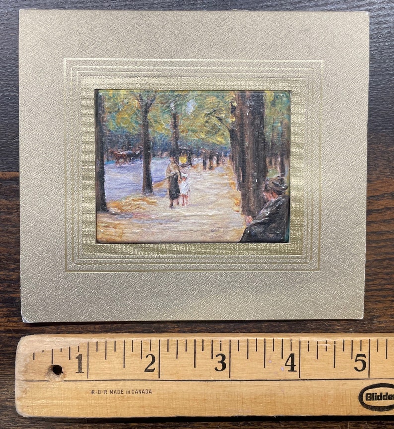 May include: A framed miniature painting depicting a park scene with figures, trees, and a pathway. The painting is set within a gold-toned frame, mounted on a textured, light-colored card. A wooden ruler with inch markings is at the bottom.