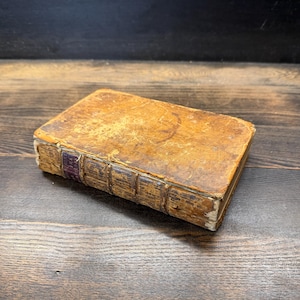 May include: An antique book with a weathered, tan leather cover and a spine featuring raised bands and a small, dark purple label. The edges of the cover show signs of wear and discoloration, with a rectangular shape and a thick, sturdy binding.