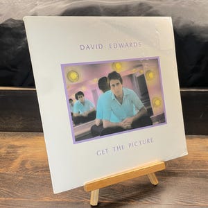 May include: A vinyl record album titled "Get The Picture" by David Edwards. The album cover features a man in a light blue shirt, seated in front of a mirror. The cover has a purple border and is displayed on a small wooden easel.