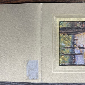 May include: A small, framed painting depicting a landscape scene with trees and water. The painting is set within a gold-colored frame, and the background is a textured, light gold color. The artwork is mounted on a folded card.