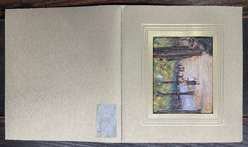 May include: A small, framed painting depicting a landscape scene with trees and water. The painting is set within a gold-colored frame, and the background is a textured, light gold color. The artwork is mounted on a folded card.