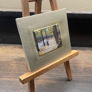 May include: A small framed painting on a wooden easel. The painting depicts a park scene with figures walking along a path. The frame is a light gold color, and the easel is made of wood.