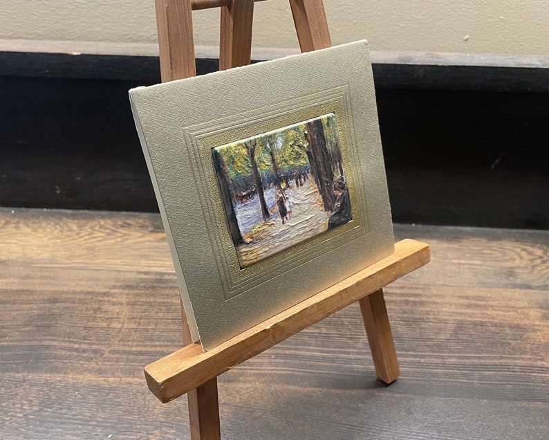 May include: A small framed painting on a wooden easel. The painting depicts a park scene with figures walking along a path. The frame is a light gold color, and the easel is made of wood.